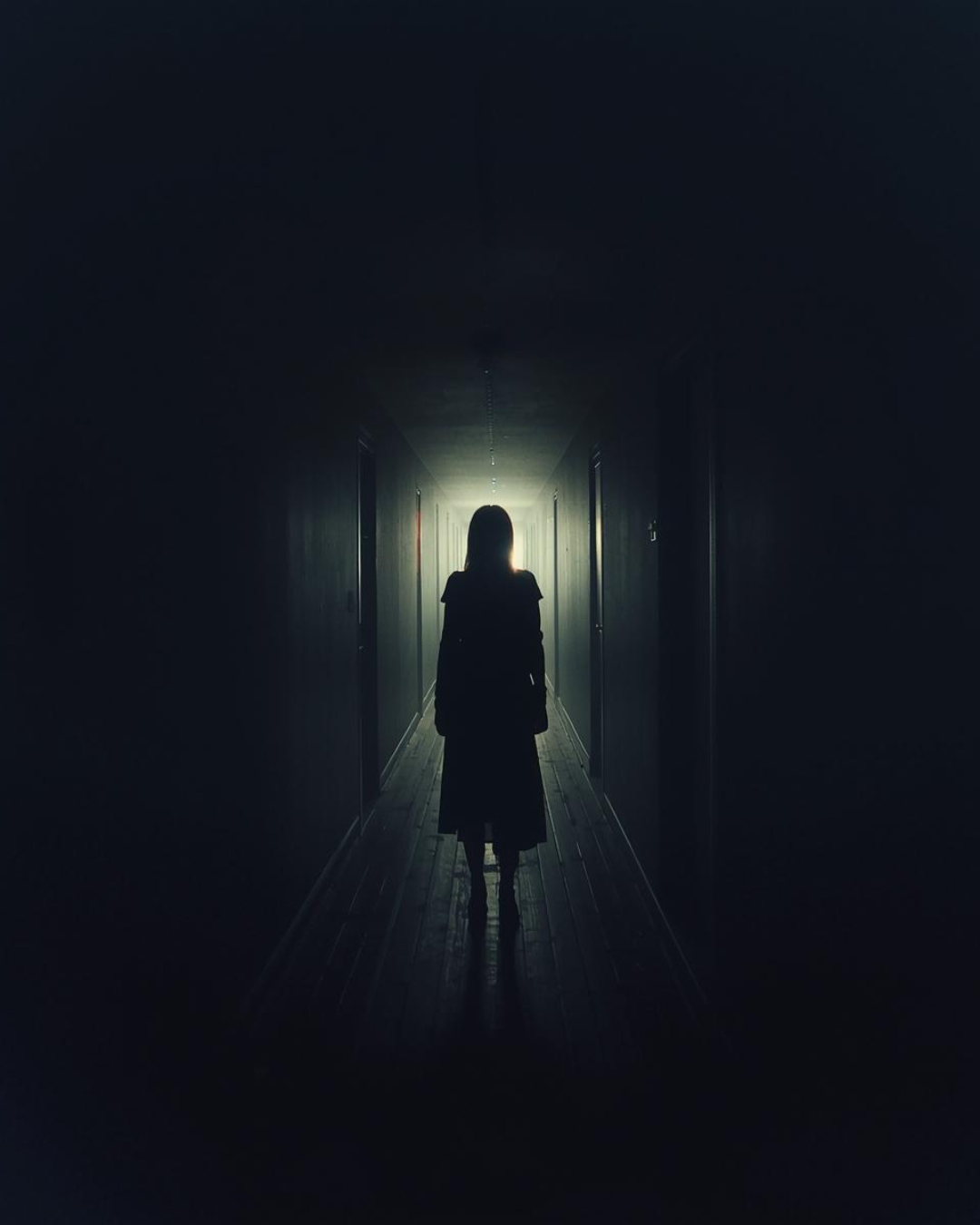 A woman in a dark hallway, walking towards light at the end.
